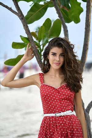 Brunette woman in red dress posing near green tree outdoorsの写真素材