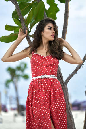 Brunette woman in red dress posing near green tree outdoorsの写真素材