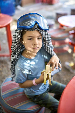Small boy in checkered keffiyeh eating banana outdoorsの写真素材
