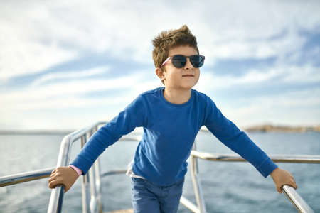 Cheerful small boy in sunglasses posing on yacht on sunny sea backgroundの写真素材