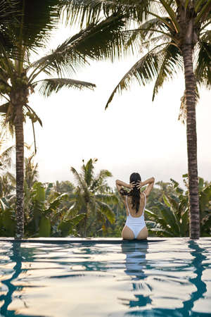 Sexy girl in a white swimsuit sits back on the edge of the outdoor swimming pool on the green jungle background on Bali. Sunlight is breaking through the clouds and reflecting in the water. Vertical.の写真素材