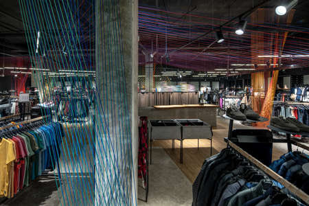 Huge clothing store with dark ceiling with hanging luminous lamps, concrete columns and multicolored ropes lines installations. There are many hangers with different clothes, shoes, cash desk.の写真素材