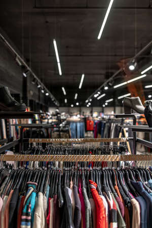 Rack with hanging colorful wear and black shelves with shoes on the blurred background of the large clothing store with many hanging luminous lamps. Closeup. Vertical.の写真素材