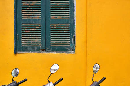 Bright yellow wall with window with wooden turquoise shutters and parked motorbikes in the front of it in Hanoi in Vietnam. Closeup. Horizontal.の写真素材