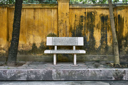 Textured granite bench between two trees on the background of the shabby yellow fence on the city street of Hanoi in Vietnam. Horizontal.の写真素材