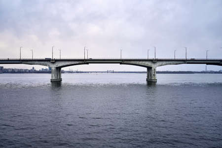 Cloudy cityscape of Dnipro with bridges through Dnieper river on the sky background in Ukraine. There are moving cars and a bicyclists on the nearest bridge. Aerial horizontal photo.の写真素材