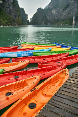 Many colorful kayaks on the wooden pier on the green rocks background in Halong Bay in Vietnam. They are orange, red, blue, yellow and green. Closeup. Vertical.の写真素材
