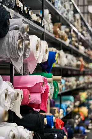 Lot of different sizes multicolored fabric rolls are lying on the gray shelves inside a warehouse....