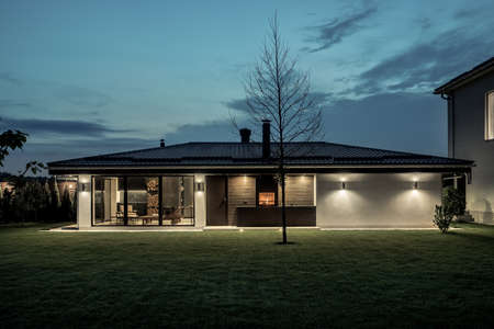Illuminated modern estate with a two storey house and a spa building with a burning fireplace on the evening sky background. It has a green lawn with trees and bushes. Horizontal.の写真素材