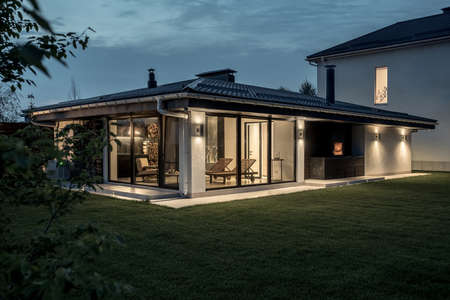 Beautiful modern estate with a two storey house and a spa building with a burning fireplace on the evening sky background. It has a green lawn with trees. Lamps are luminous. Horizontal.の写真素材