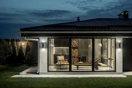 Luminous modern spa building with a glass wall with doors on the dark sky background. Inside it there are deck chairs, armchair, table, stand with firewood. There is green lawn with trees and bushes.の写真素材
