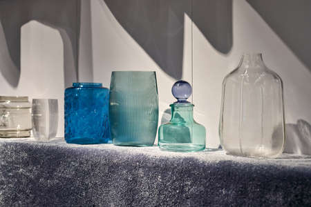 Different multicolored glass vases on the textured soft surface on the light wall background in the illuminated interior. Closeup. Horizontal.の写真素材
