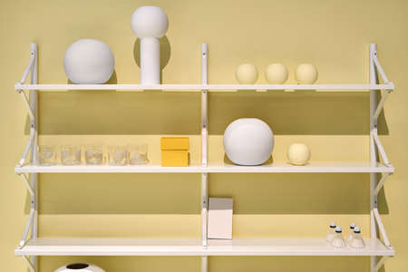 White shelves on the yellow wall in the illuminated interior. On them there are different vases,...