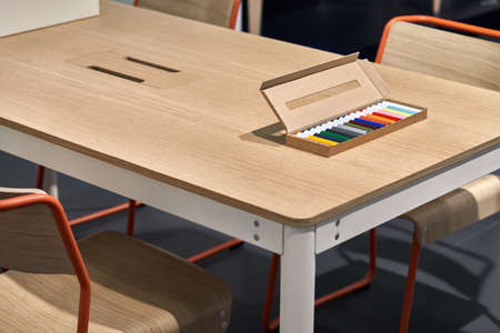 Cardboard box with multicolored markers lies on the wooden table in the illuminated interior. There...