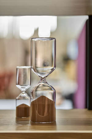 Couple of sandglasses on the light wooden table on the blurred background indoors. Closeup vertical...