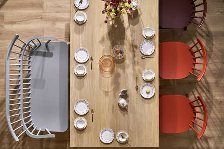 Wooden table with white dishes with flower prints, colorful chairs and gray sofa in the luminous...