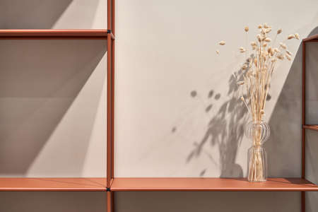 Metal orange shelves with a glass vase with dried plants on the light wall background in the...