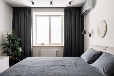 Contemporary bedroom with white walls with a relief picture. There is a double bed with gray linens and pillows, black hanging lamp, window with dark curtains, clock, green plant in the pot, stand.の写真素材