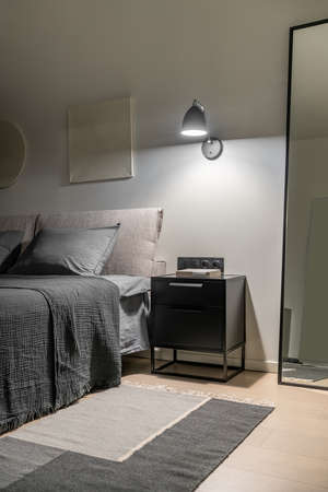 Amazing modern bedroom with white walls with relief pictures and a parquet with a multicolored carpet. There is a double bed with gray linens and pillows, black nightstand, luminous lamp, tall mirror.の写真素材