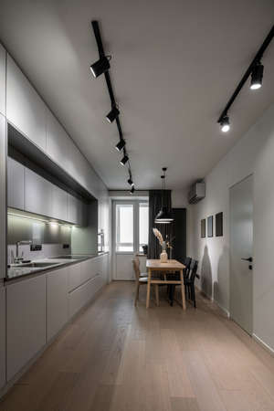 Modern kitchen with light walls and a parquet. There are gray lockers, sink with chrome faucet, stove, oven, wooden table with vase and multicolored chairs, window with curtain, luminous lamps, door.の写真素材