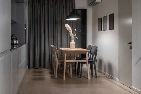 Contemporary kitchen with light walls and a parquet floor. There are gray lockers, stove with a chrome kettle, oven, wooden table with vase and chairs, dark curtains, hanging luminous lamps, door.の写真素材