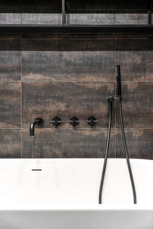 Stylish illuminated modern bathroom with textured bronze tiled walls. There is a white bath with a dark matte faucet and shower, black shelves. Closeup. Vertical.の写真素材