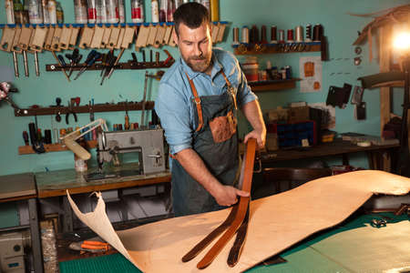 Craftsman in apron working with leather at workshop, belt manufacturing process.