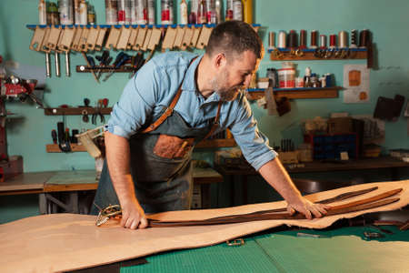 Craftsman in apron working with leather at workshop, belt manufacturing process.の写真素材