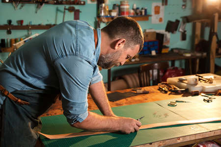 Craftsman in apron working with leather at workshop, belt manufacturing process.の写真素材