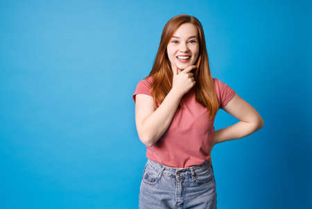 Thinking and smiling beautiful young woman in pink t-shirt and jeans posing isolated on blue wall background.の写真素材