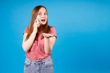 Girl with surprised expression talking at the phone. Ginger girl in pink t-shirt and jeans isolated on blue backgroundの写真素材