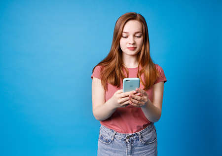 Young ginger girl in pink t-shirt and jeans posing isolated on blue background. Using mobile phone, typing sms message.の写真素材