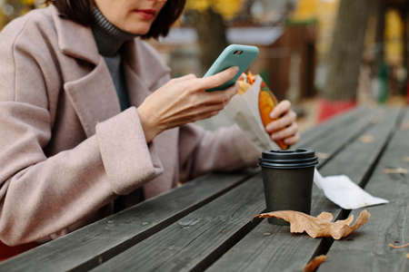 woman eating hot dog in autumn park and drinking coffee sitting at old wooden tableの写真素材
