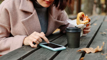 woman eating hot dog in autumn park and drinking coffee sitting at old wooden tableの写真素材