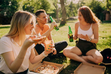 Young blonde girl in sunglassesdrinking alcohol and eating pizza with her friends outdoorsの写真素材