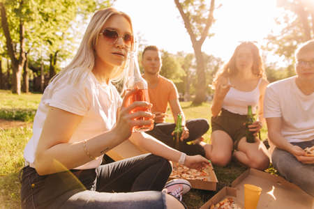 Young blonde girl in sunglassesdrinking alcohol and eating pizza with her friends outdoorsの写真素材