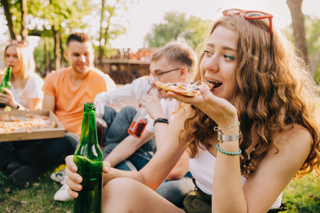 Young curly girl eating pizza and drinking beer together with her friends outdoorsの写真素材
