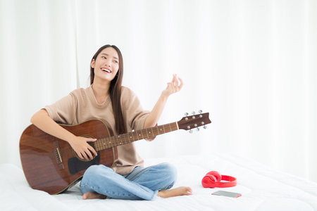 Beautiful Asian woman is smiling with playing guitar  on the bedの写真素材