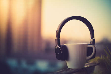 Headphones and coffee cup on blurred city background. Music concept. View of sunrise time, Vintage...