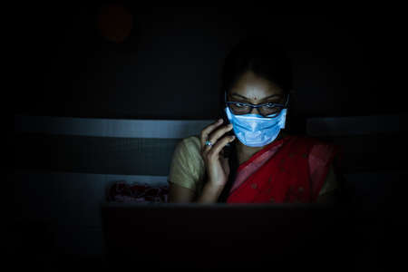 Young indian Woman wearing mask working on her laptop and talking on phone, late night working,...