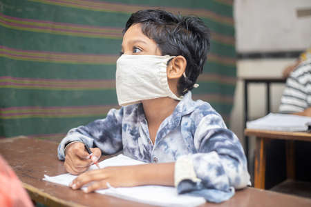 Indian boys students studying In classroom wearing mask maintaining social distancing looks at...