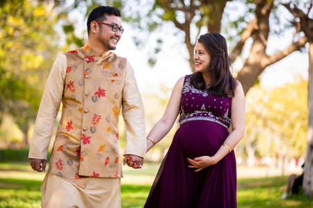 Happy asian indian pregnant woman with her husband in walking outdoor in a park or garden, smiling cheerful young parents looking at camera expecting baby.の写真素材