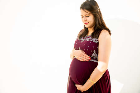 Young asian indian pregnant woman looking at belly standing against white background outdoor studio shot, happy female expecting baby, motherhood and pregnancy concept, copy space.の写真素材