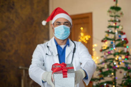 Doctor wearing mask and santa hat showing christmas gift box, celebration during covid-19 pandemic,...