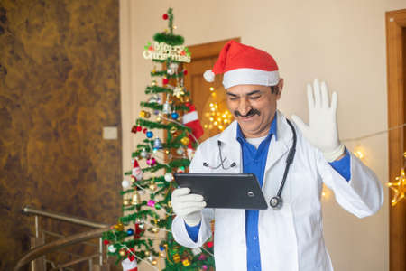 Male middle age doctor wearing santa hat video calling over tablet, christmas celebration during...