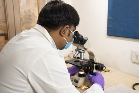 Asian Indian male medical doctor or researcher wearing mask looking through a microscope in a...