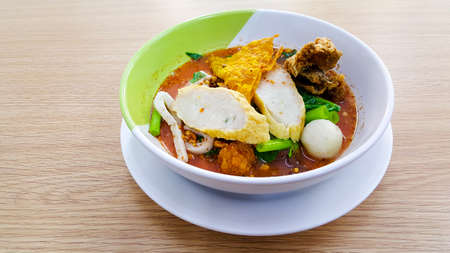 Noodle bowl that consist of fish ball, vegetable, soup and more placed on the white cup and disk on the wooden table.の写真素材