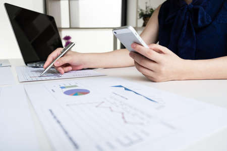 Business women is holding mobile phone and analyzing marketing strategy from the selling performance graph in the office room, Finance and accounting concept.の写真素材