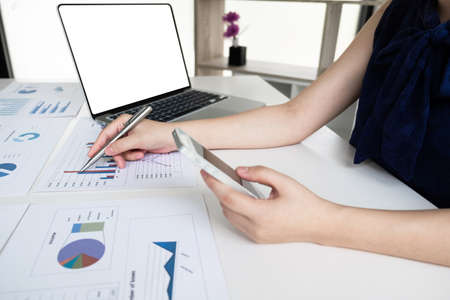 Business women is holding mobile phone and analyzing marketing strategy from the selling performance graph in the office room, Finance and accounting concept.の写真素材