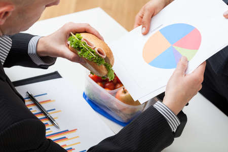 Businessman with pie chart eating sandwich during workの写真素材